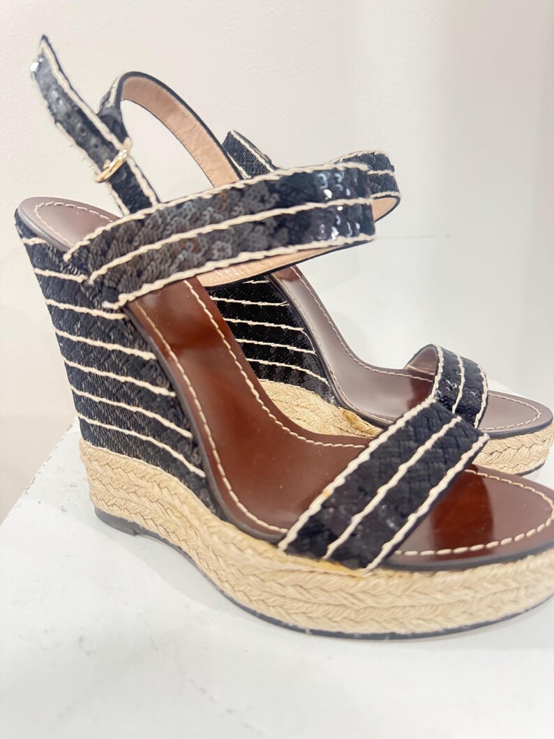 May include: A pair of black wedge sandals with white striped detailing. The sandals have a woven jute platform and a brown leather insole. The straps are black with sequin accents and a gold buckle.