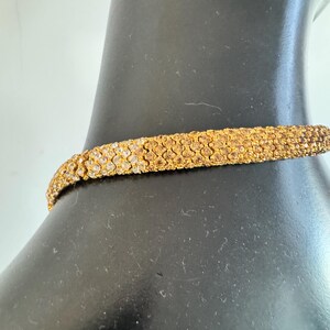 K&ouml;nnte beinhalten: Ein goldfarbenes Choker-Halsband mit strukturierter Oberfl&auml;che. Die Halskette besteht aus kleinen, eng aneinander liegenden Elementen, die ein detailliertes Muster bilden. Der Schmuck wird auf einer schwarzen Schaufensterpuppe pr&auml;sentiert.