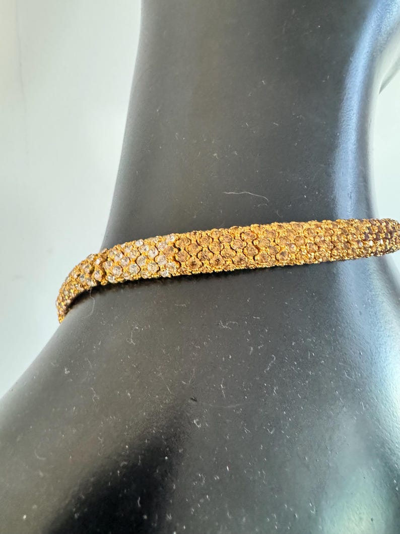 K&ouml;nnte beinhalten: Ein goldfarbenes Choker-Halsband mit strukturierter Oberfl&auml;che. Die Halskette besteht aus kleinen, eng aneinander liegenden Elementen, die ein detailliertes Muster bilden. Der Schmuck wird auf einer schwarzen Schaufensterpuppe pr&auml;sentiert.