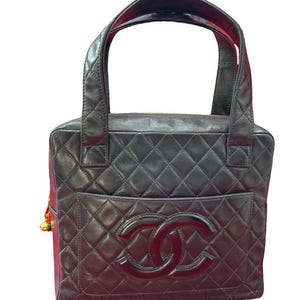 May include: Black quilted leather tote bag with a large front pocket and a prominent double-C logo. The bag features two top handles and a gold-toned charm. The interior lining is red. The bag has a rectangular shape.