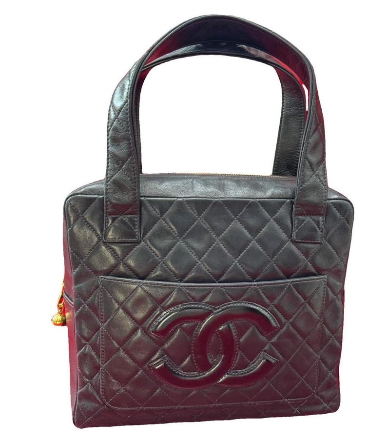 May include: Black quilted leather tote bag with a large front pocket and a prominent double-C logo. The bag features two top handles and a gold-toned charm. The interior lining is red. The bag has a rectangular shape.