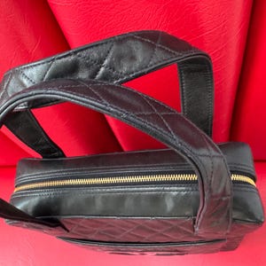 May include: Black leather handbag with a quilted pattern and a gold zipper. The bag has a top handle and a rectangular shape. The bag is sitting on a red surface.