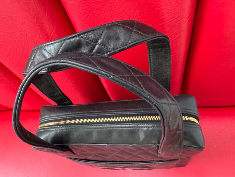 May include: Black leather handbag with a quilted pattern and a gold zipper. The bag has a top handle and a rectangular shape. The bag is sitting on a red surface.
