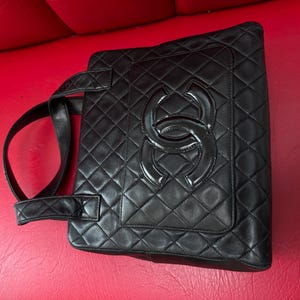 May include: Black quilted leather tote bag with a large, glossy interlocking logo on the front. The bag features a diamond pattern and two top handles. The bag is set against a red background.