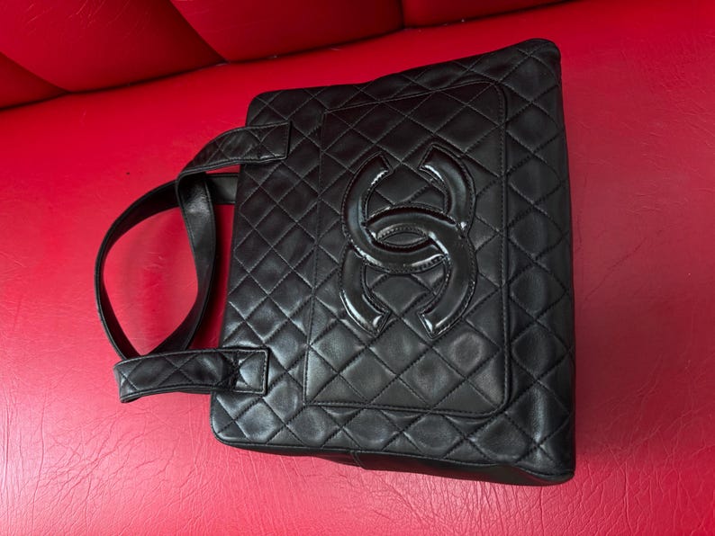 May include: Black quilted leather tote bag with a large, glossy interlocking logo on the front. The bag features a diamond pattern and two top handles. The bag is set against a red background.