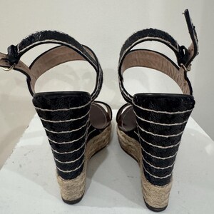 May include: Pair of black wedge sandals with a woven jute sole and ankle straps. The wedge heels are adorned with horizontal black and white stripes. The sandals have a buckle closure and a platform sole.
