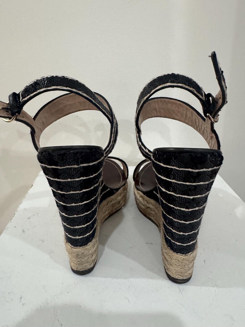 May include: Pair of black wedge sandals with a woven jute sole and ankle straps. The wedge heels are adorned with horizontal black and white stripes. The sandals have a buckle closure and a platform sole.