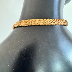 K&ouml;nnte beinhalten: Eine goldfarbene Halskette mit Perlen. Die Halskette besteht aus kleinen, eng gewebten Perlen, die eine strukturierte Oberfl&auml;che bilden. Der Choker wird auf einer schwarzen Schaufensterpuppe pr&auml;sentiert, die sein Design und seine Farbe zeigt.