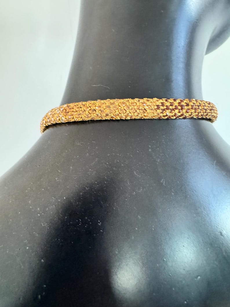 K&ouml;nnte beinhalten: Eine goldfarbene Halskette mit Perlen. Die Halskette besteht aus kleinen, eng gewebten Perlen, die eine strukturierte Oberfl&auml;che bilden. Der Choker wird auf einer schwarzen Schaufensterpuppe pr&auml;sentiert, die sein Design und seine Farbe zeigt.