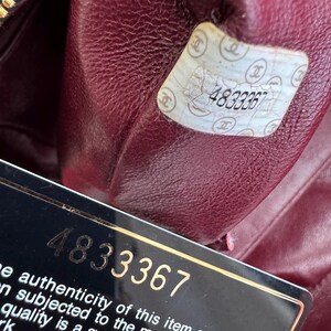 May include: Close-up of the interior of a black leather handbag with a burgundy lining. A white sticker with the number "4833367" is visible, along with a black card bearing the same number and text about authenticity.