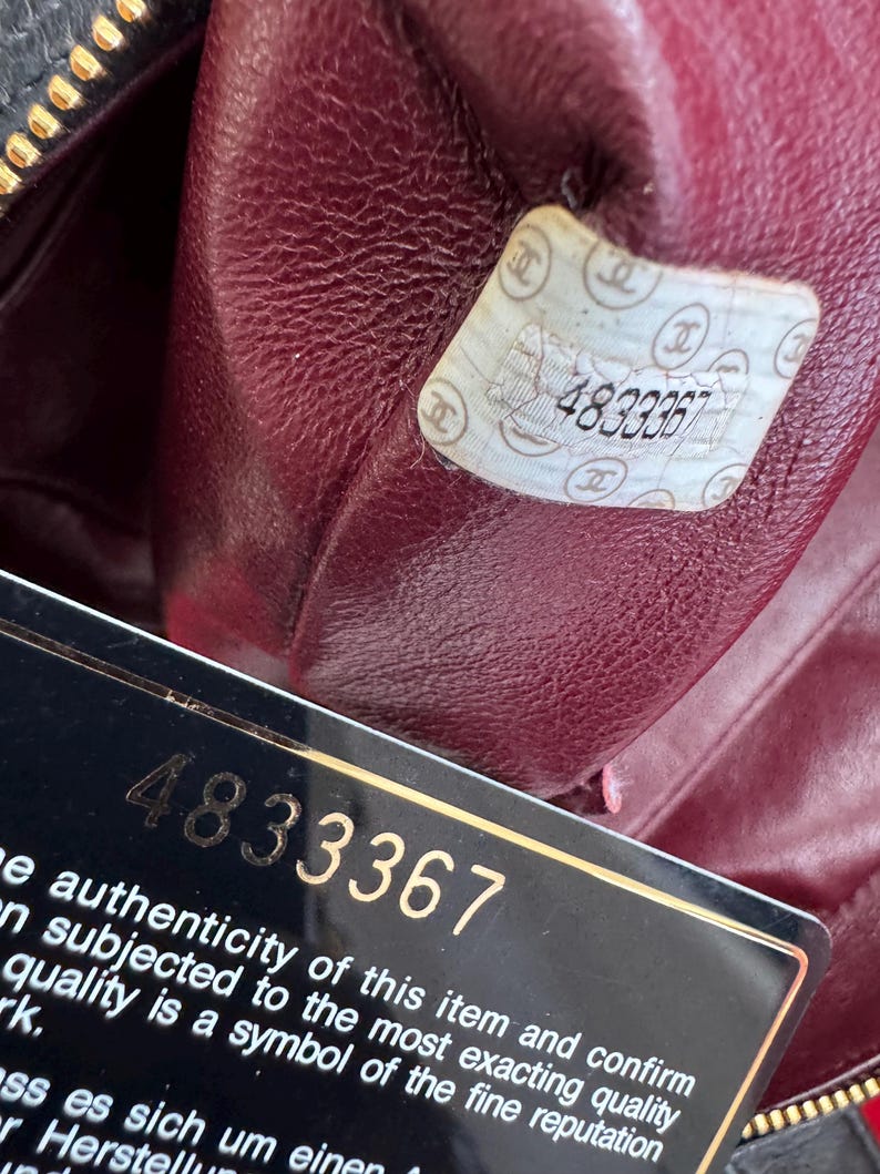 May include: Close-up of the interior of a black leather handbag with a burgundy lining. A white sticker with the number "4833367" is visible, along with a black card bearing the same number and text about authenticity.
