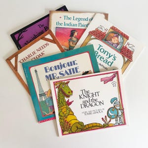 May include: A collection of children's books with colorful covers. Titles include "The Knight and the Dragon," "Bonjour, Mr. Satie," and "Tony's Bread." The books are arranged in a fan shape, showcasing their illustrations and titles.
