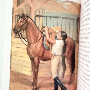 May include: A watercolor illustration of a brown horse standing in a wooden stable. A person with dark hair, wearing a white shirt, beige breeches, and black riding boots, is placing a tan leather saddle onto the horse's back.