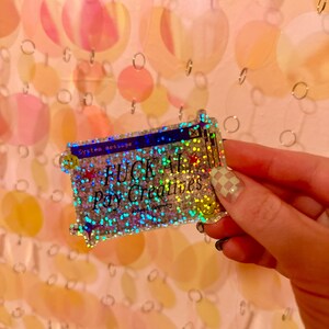 May include: A rectangular holographic sticker with a glittery finish featuring a dark blue computer window design. The text reads System message, FUCK AI, Pay Creatives, and @doodlymeg, accented with small red stars and yellow smiley faces.