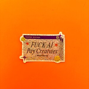 May include: A rectangular holographic sticker featuring a purple header with the text System message and an X icon. The center text reads FUCK AI Pay Creatives in black, flanked by red stars. Two yellow smiley faces and purple star icons decorate the corners.