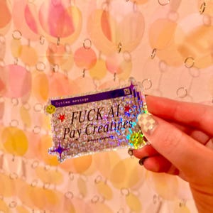 May include: A rectangular holographic sticker featuring a purple computer window frame with the text "System message" and "FUCK AI Pay Creatives" in black. The design includes small red stars, purple sparkles, and yellow smiley faces.