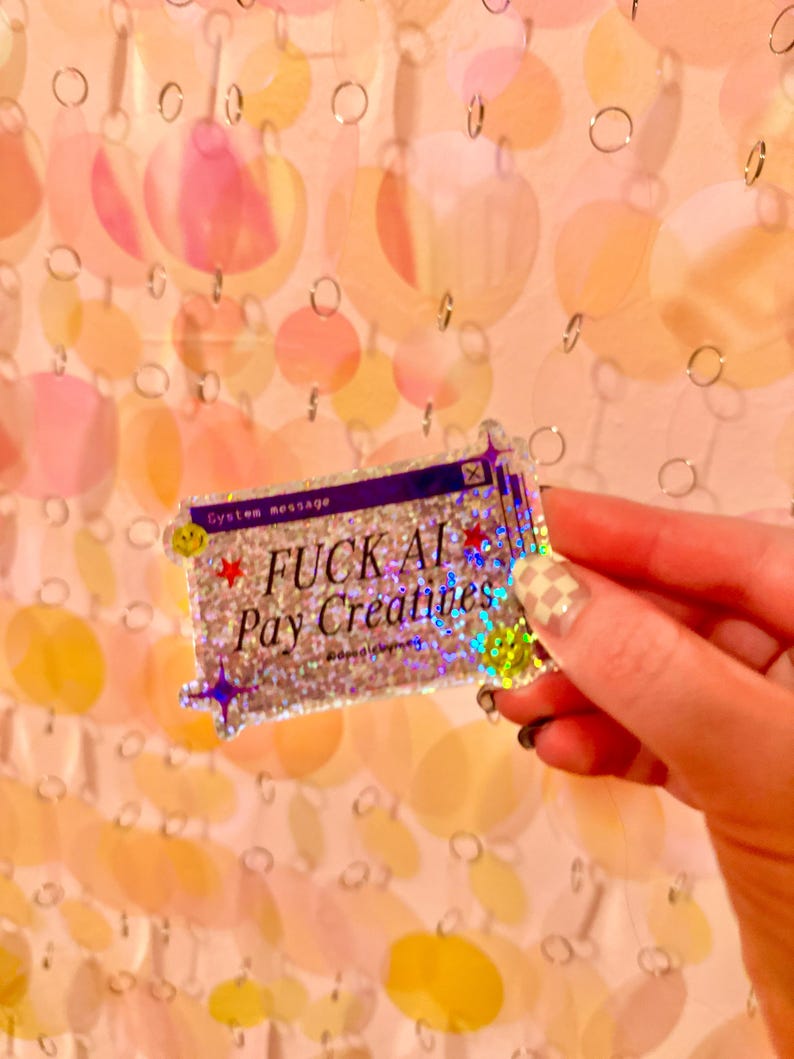 May include: A rectangular holographic sticker featuring a purple computer window frame with the text "System message" and "FUCK AI Pay Creatives" in black. The design includes small red stars, purple sparkles, and yellow smiley faces.