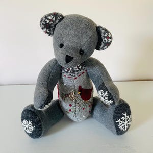 May include: A gray teddy bear with a winter-themed sweater and snowflake details on its feet. The sweater features a red cardinal, pine cones, and holly.