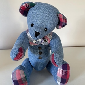 May include: A blue denim teddy bear with plaid fabric accents. The bear has black button eyes and a plaid bow tie.