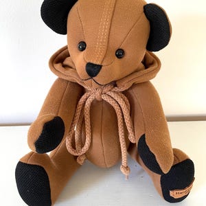 May include: A handmade brown teddy bear with black accents on the ears, paws, and nose. The bear wears a matching hoodie with a drawstring. The bear has black button eyes and a stitched mouth. A small tag reads "Handmade".
