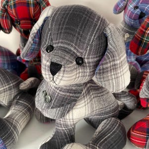 May include: A collection of handmade plush dog toys in various plaid patterns. The central dog is gray and white, with black button eyes and a black nose. Other dogs in the image feature red, blue, and other plaid fabrics. Each dog has button details.