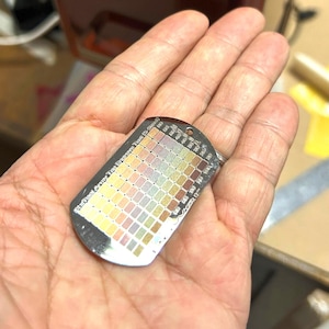 Puede incluir: Un colgante de placa de identificación metálica con una cuadrícula de pequeños cuadrados multicolores dispuestos en filas y columnas. La superficie muestra un degradado de tonos que van del amarillo y naranja al rosa, verde y marrón, con texto grabado.