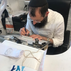 May include: A person wearing a black yarmulke and white shirt is writing on a white clipboard. A small white bag with a brown string is on a white surface with a blue and white surfboard.