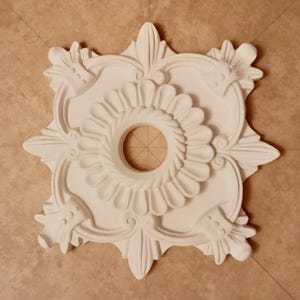 Puede incluir: Medallón de techo de yeso blanco con un diseño floral y una abertura circular central. El medallón tiene un diseño simétrico con cuatro puntos.