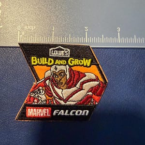 May include: A black, orange, and red patch featuring the Marvel character Falcon. The patch includes the text "LOWE'S BUILD AND GROW" and "MARVEL FALCON". The Falcon is depicted in his red and white costume.