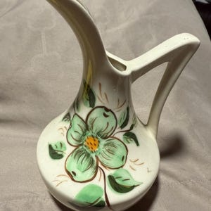 May include: A white ceramic pitcher with a green floral design. The pitcher has a long, curved spout and a handle.