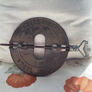 May include: A vintage, rusty metal reversible spindle with a wire handle. The text "REVERSIBLE" and "STEEL 7 IN SPINDLE" are embossed on the circular piece. The handle is made of coiled wire.