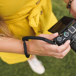 May include: A person holding a black Canon camera with a flip-out screen. The camera has a lens with a silver ring and a black strap.