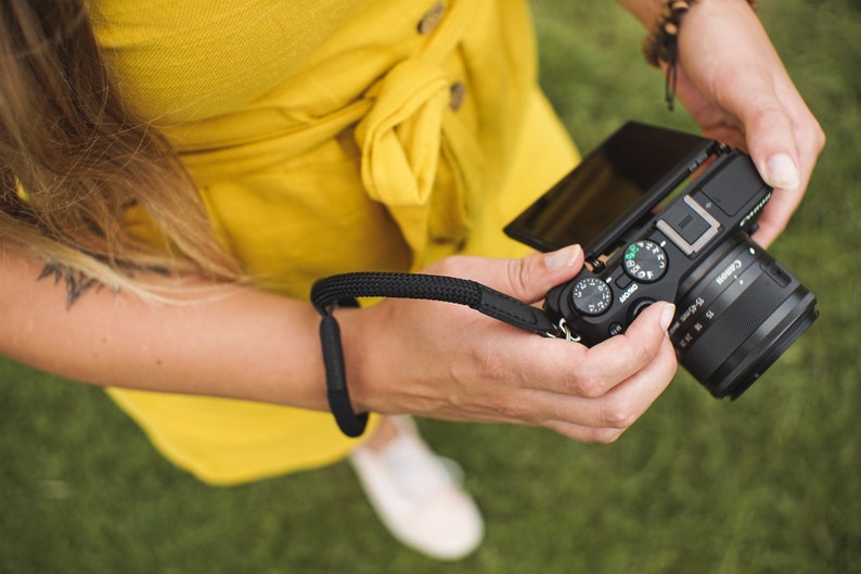 May include: A person holding a black Canon camera with a flip-out screen. The camera has a lens with a silver ring and a black strap.