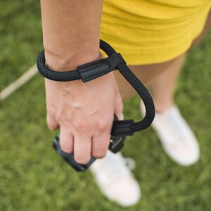 May include: A black camera wrist strap with a leather patch. The strap is worn around a person's wrist and is attached to a camera.