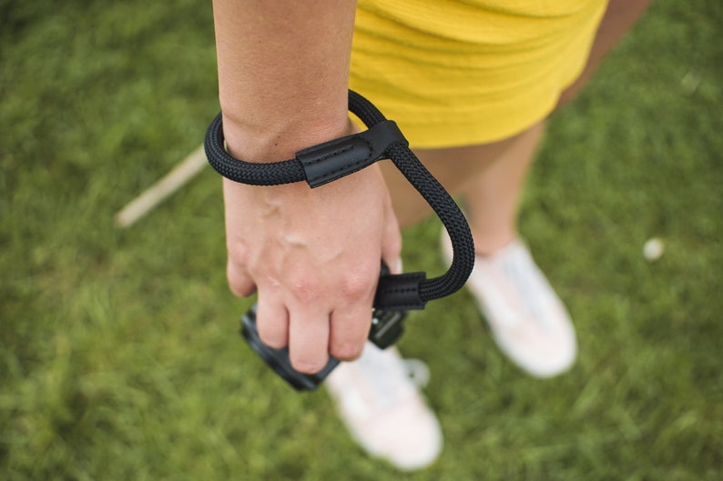 May include: A black camera wrist strap with a leather patch. The strap is worn around a person's wrist and is attached to a camera.