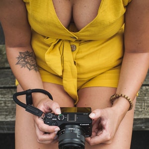 May include: A person wearing a yellow jumpsuit holds a black Canon camera with a lens. The camera has a black strap attached to it.