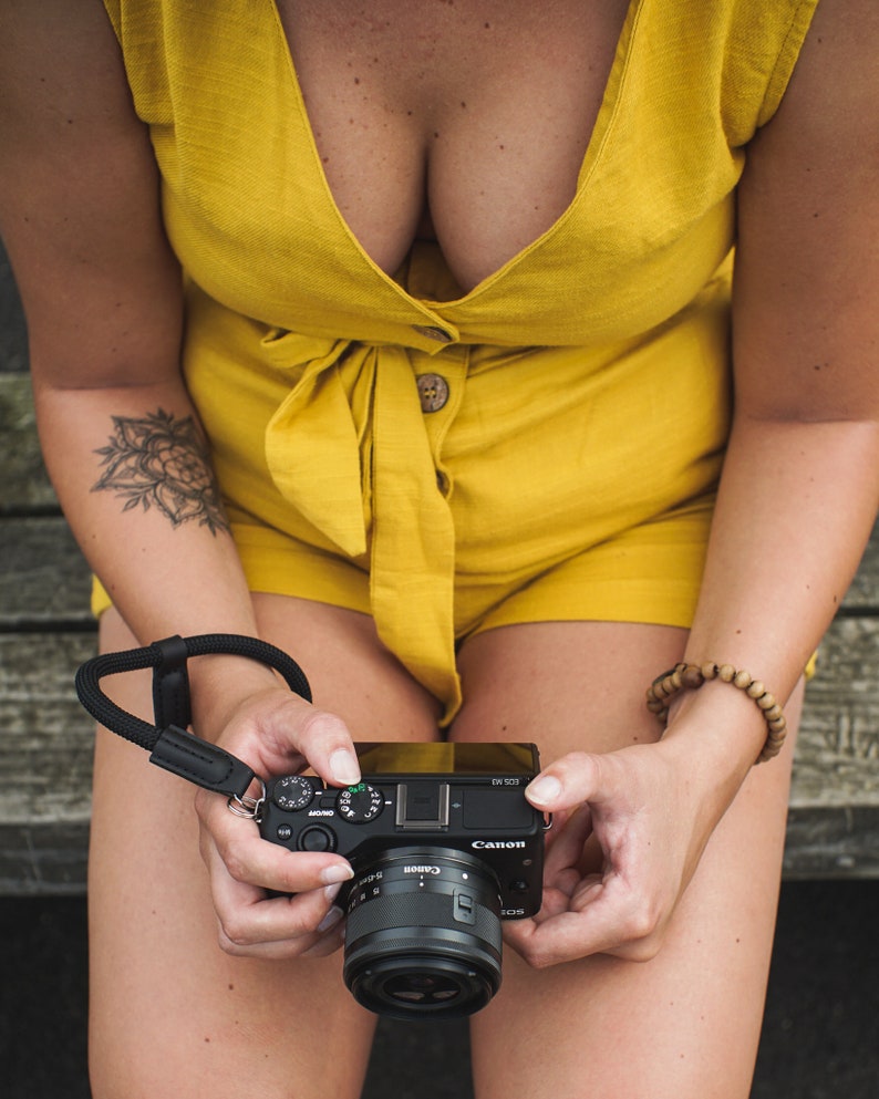 May include: A person wearing a yellow jumpsuit holds a black Canon camera with a lens. The camera has a black strap attached to it.