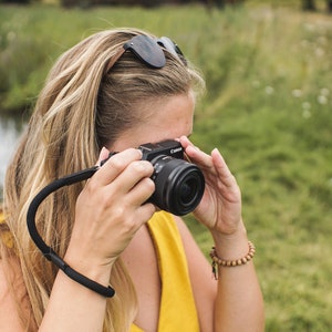 May include: A woman in a yellow dress is holding a black camera with a black strap. She is looking through the viewfinder of the camera.