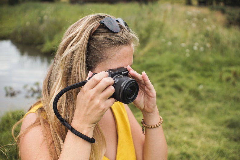May include: A woman in a yellow dress is holding a black camera with a black strap. She is looking through the viewfinder of the camera.