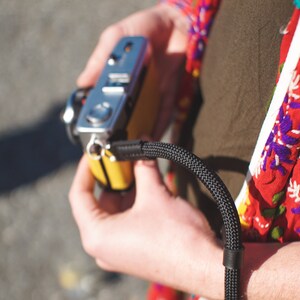 May include: A person holding a silver and yellow camera with a black camera strap. The camera strap is made of a woven material.