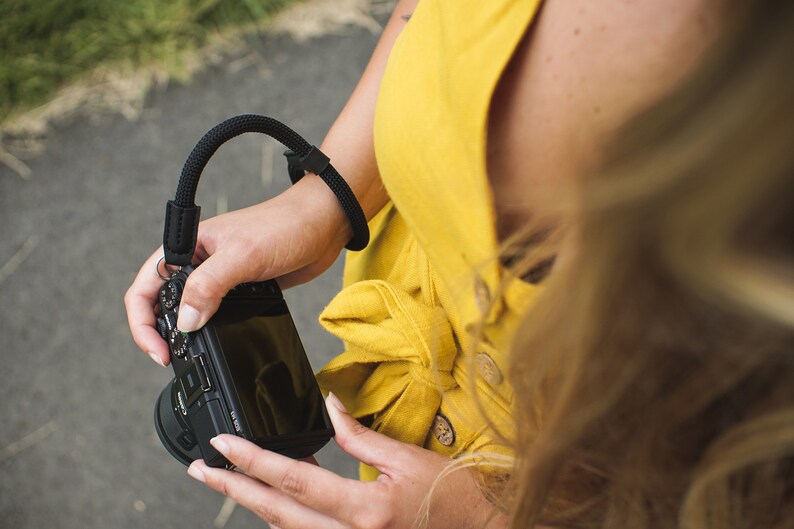 May include: A person wearing a yellow dress holds a black camera with a black wrist strap. The camera has a black lens and a screen on the back.