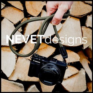 May include: A black Canon camera with a green and black camera strap. The camera strap is held by a hand against a background of stacked wooden logs. The text "NEVET designs" is visible in the image.