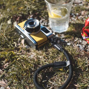May include: A yellow and silver camera with a black camera strap lying on green grass. A glass of water with a lemon wedge is in the background.