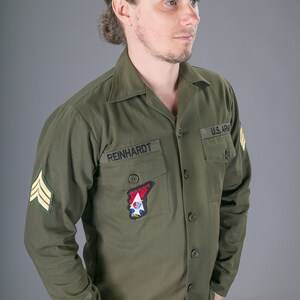 May include: A man wearing a green long-sleeved shirt with a U.S. Army patch, a patch that says "Allen Scouts", and a name tag that says "REINHARDT".
