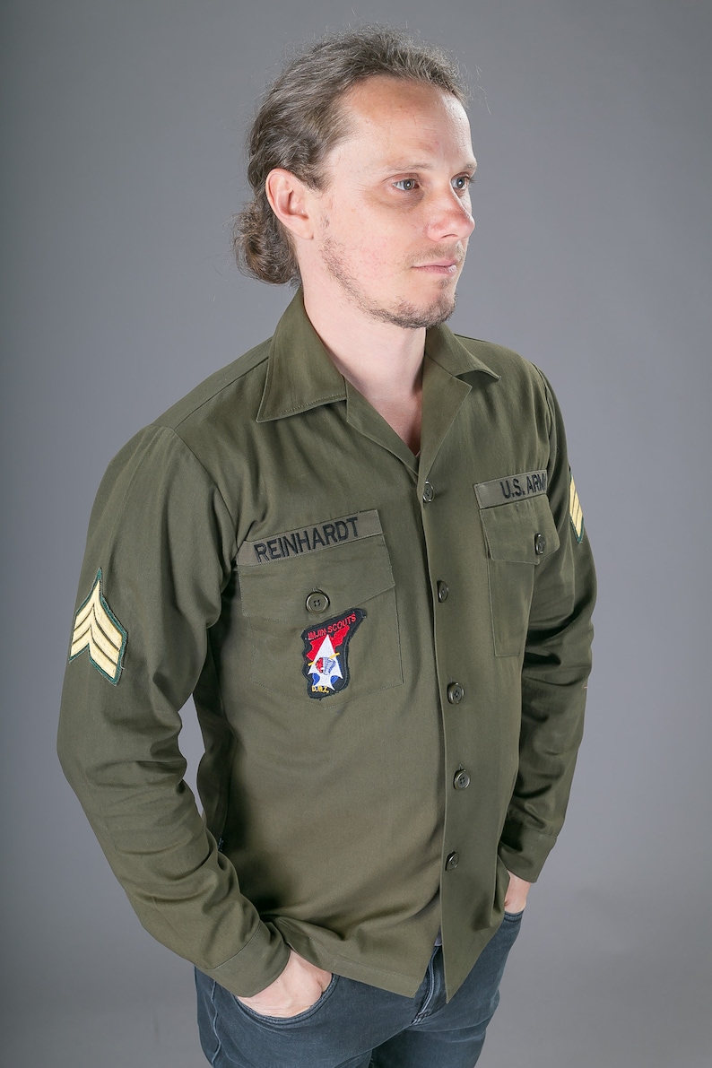 May include: A man wearing a green long-sleeved shirt with a U.S. Army patch, a patch that says "Allen Scouts", and a name tag that says "REINHARDT".