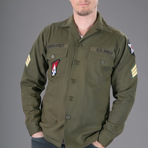 May include: A green long-sleeved shirt with a button-up front and two pockets. The shirt has a patch on the left pocket that says "REINHARDT" and a patch on the right pocket that says "U.S. ARMY".