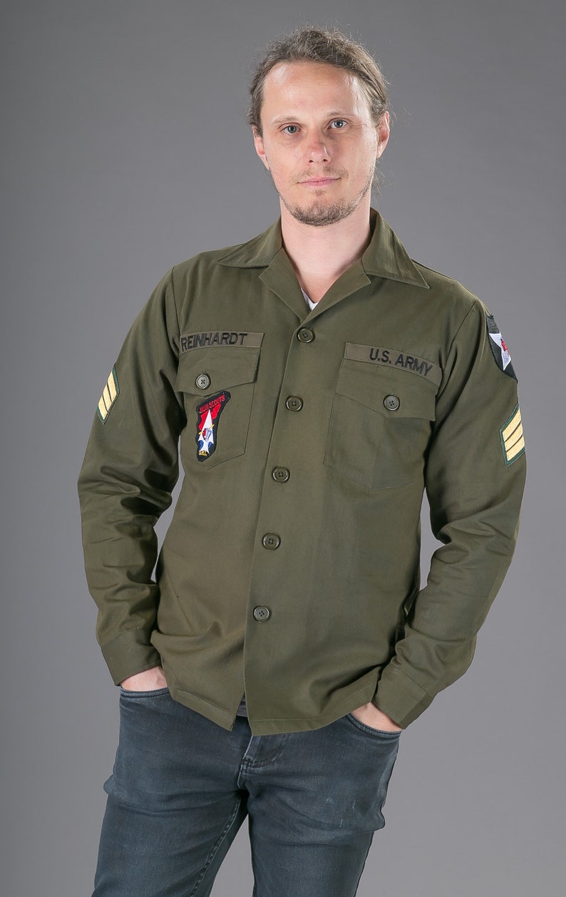 May include: A green long-sleeved shirt with a button-up front and two pockets. The shirt has a patch on the left pocket that says "REINHARDT" and a patch on the right pocket that says "U.S. ARMY".