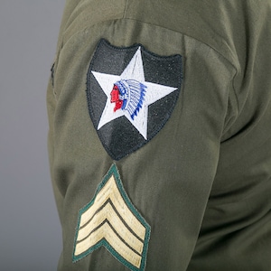 May include: A green military uniform with a black and white patch featuring a star and an American Indian head. The patch is on the left sleeve of the uniform. There is also a gold chevron patch on the sleeve.