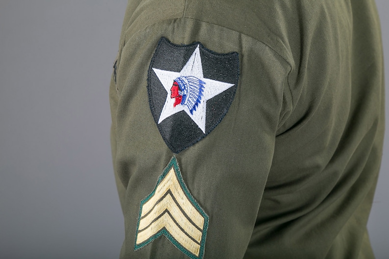 May include: A green military uniform with a black and white patch featuring a star and an American Indian head. The patch is on the left sleeve of the uniform. There is also a gold chevron patch on the sleeve.