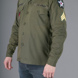 May include: A green long-sleeved shirt with a button-up collar and two pockets. The shirt has the text "US ARMY" on the left pocket and a star patch on the right sleeve.